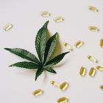 Flat lay of cannabis leaf surrounded by gel capsules on a white surface.